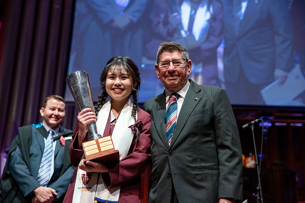 Sam To on stage at Speech Night, holding the Cameron Cup beside a College benefactor