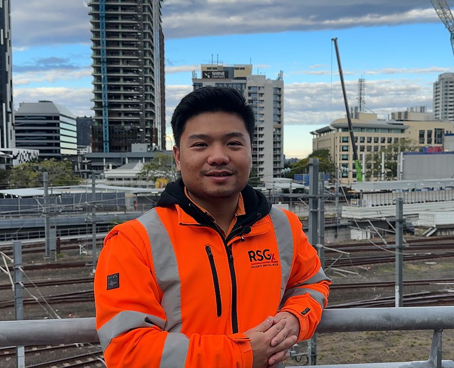JPC alumnus Joseph Vu in an orange hi-vis jacket at the Cross River Rail site, Brisbane skyline behind him