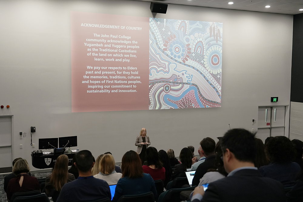 A presenter at the lectern beside the Acknowledgement of Country slide, with delegates seated in the lecture theatre