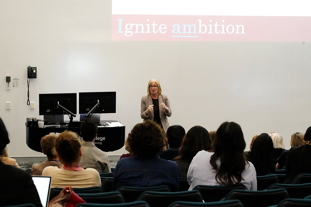 A presenter speaks to delegates in the lecture theatre beneath a slide reading Ignite ambition