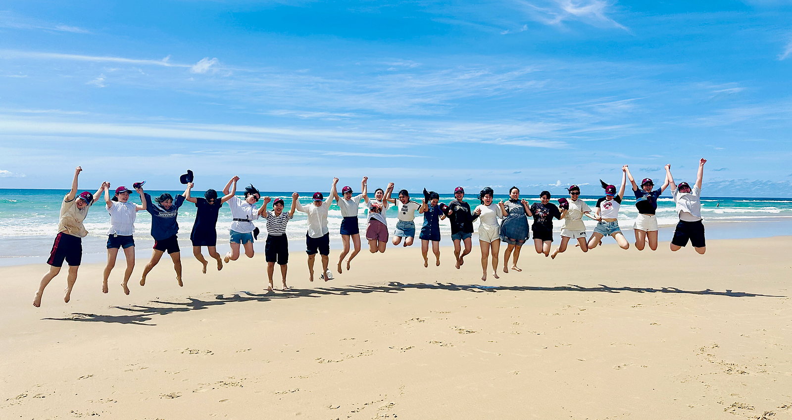 International students jumping in the air together on a wide white-sand Queensland beach with turquoise water behind them