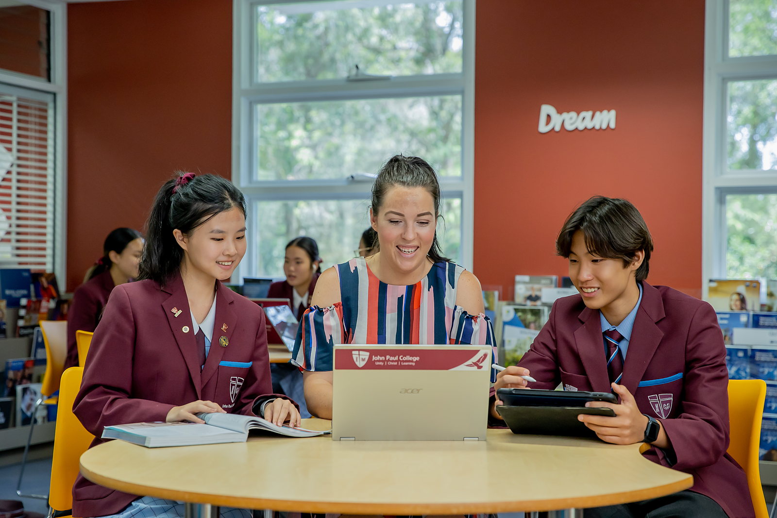 A John Paul College teacher working at a library table alongside two international students, all three looking at a laptop together